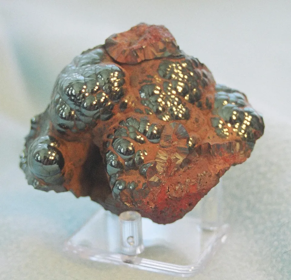 A rounded lump of hematite, shiny black-gray on top and rust-red underneath, on a clear stand.